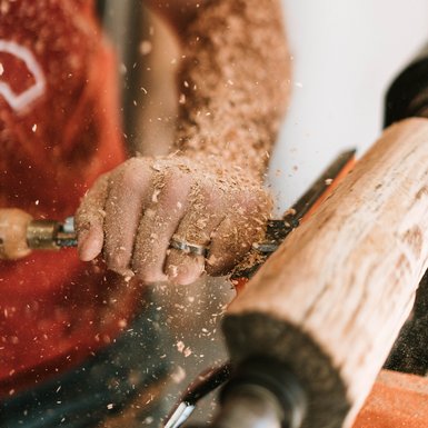 Nahaufnahme der Hand einer Person, die mit einem Hobel ein stück Holz auf einer Drehbank bearbeitet.