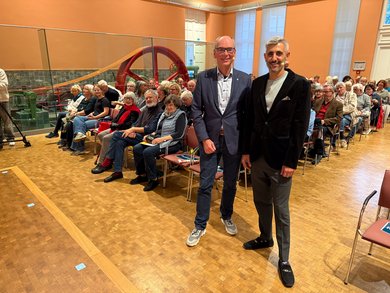 Bild: VHS Oldenburg VHS-Geschäftsführer Andreas Gögel und Michel Abdollahi stehn vor dem Publikum und lächeln in die Kamera.