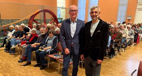 VHS-Geschäftsführer Andreas Gögel und Michel Abdollahi stehn vor dem Publikum und lächeln in die Kamera.