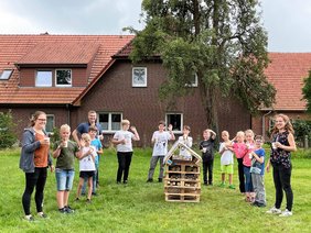 12 Kinder und Jugendliche, Dr. Christian Pundt und die Dozentin stehen mit Getränken im Halbkreis auf einer Wiese. In der Mitte steht ein ca. 1 Meter hohes Insektenhotel aus Paletten und anderem Holz. Auf einem Schild am Insektenhotel steht "Insektenvilla Kunterbunt". Bild: Uta Wilms,Gemeinde Hatten.