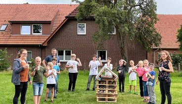 12 Kinder und Jugendliche, Dr. Christian Pundt und die Dozentin stehen mit Getränken im Halbkreis auf einer Wiese. In der Mitte steht ein ca. 1 Meter hohes Insektenhotel aus Paletten und anderem Holz. Auf einem Schild am Insektenhotel steht "Insektenvilla Kunterbunt". Bild: Uta Wilms,Gemeinde Hatten.