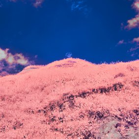 Eine Landschaft in verfremdeten Farben: Ein rosaner Hügel vor einem tiefblauen Himmel mit rosanen Wolken..