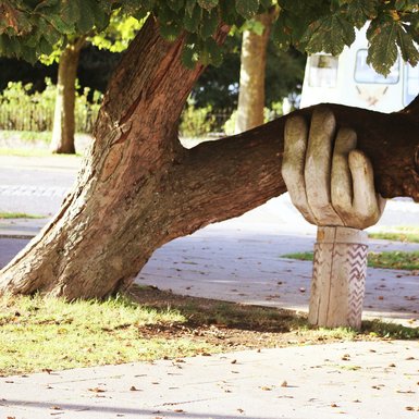 Eine Holzskulptur in Form einer Hand stützt den Ast eines Baumes.