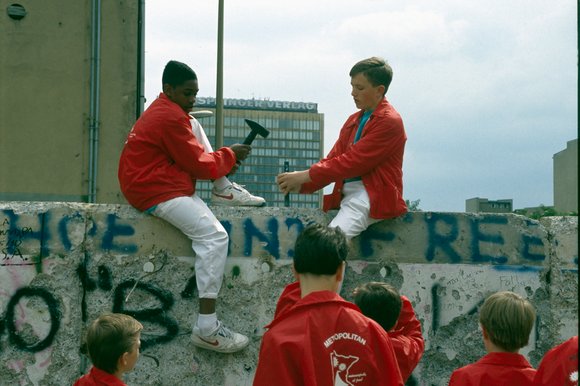Bild: Eastworldblock Jungen Menschen in roten Jacken stehen vor der Berliner Mauer. Zwei sitzen auf ihr und bearbeiten sie mit Hämmern.