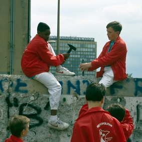 Bild: Bundesstiftung Aufarbeitung, Eastblockworld Jungen Menschen in roten Jacken stehen vor der Berliner Mauer. Zwei sitzen auf ihr und bearbeiten sie mit Hämmern.