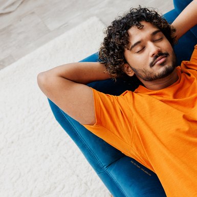 Ein junger Mann im liegt mit geschlossenen Augen und den Armen hinter seinem Kopf verschränkt auf einem blauen Sofa. Er trägt ein orangenes T-Shirt. Er hat dunkle lockige Haare, einen leichten Bart und braune Haut.