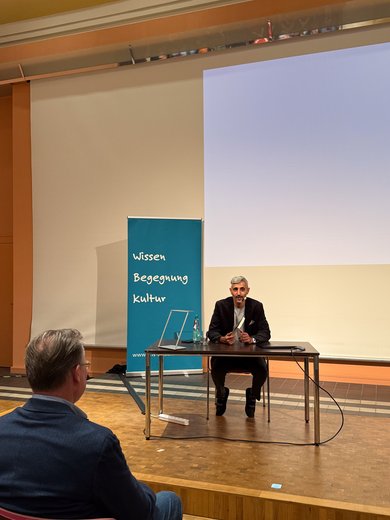 Bild: VHS Oldenburg Michel Abdollahi sitzt auf der Bühne an einem Tisch und schaut in das Publikum.
