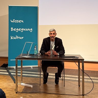 Bild: VHS Oldenburg Michel Abdollahi sitzt auf der Bühne an einem Tisch und schaut in das Publikum.