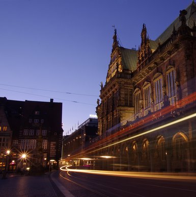 Blick auf das Bermer Rathaus bei Dunkelheit. Im Vordergrund Lichtstreifen vorbeifahrender Straßenbahn.
