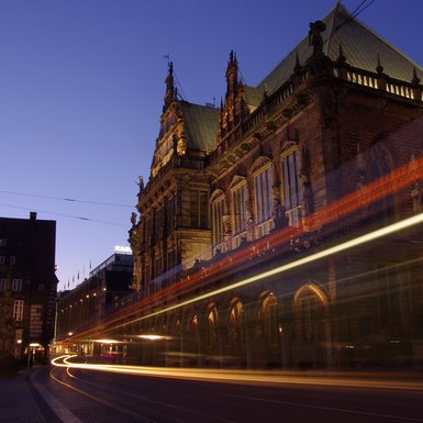 Blick auf das Bermer Rathaus bei Dunkelheit. Im Vordergrund Lichtstreifen vorbeifahrender Straßenbahn.