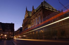 Blick auf das Bermer Rathaus bei Dunkelheit. Im Vordergrund Lichtstreifen vorbeifahrender Straßenbahn.