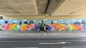 Das Wandbild in voller Länge entlang der Brückenwand. Es zeigt eine Silhuette von Oldenburg im Hintergrund. Der Schriftzug Oldenburg bleibt bunt ist in vielen bunten Farben mit Klecksen, Sternen und anderen Formen verziert und durch Sprühdpsen mit Augen und Armen und Beinen ergänzt. Bild: VHS Oldenburg