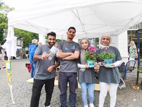 Zwei Teilnehmer und zwei Teilnehmerinnen der Oldenburger Jugendwerkstatt vor dem Stand der Jugendwerkstatt halten Blumen in die Kamera und lächeln. Bild: VHS Oldenburg.