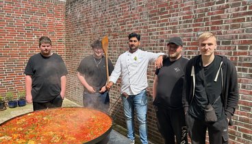 Um eine sehr große Pfanne mit gebratenem Gemüse stehen einige junge Männer. Einer von ihnen hält einen großen Kochlöffel in der Hand.