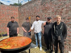 Um eine sehr große Pfanne mit gebratenem Gemüse stehen einige junge Männer. Einer von ihnen hält einen großen Kochlöffel in der Hand.