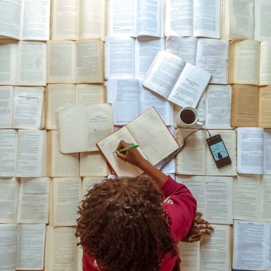 Blick von oben: Auf dem Boden ind das ganze Bild füllend Bücher aufgeschlagen. Auf den Büchern liegt eine Frau mit dunklen Locken und schreibt in ein Buch. Neben ihr stehen eine Tasse Kaffe und liegt ein Smartphone. Titel: M von Emmanuel Adegbenro.ay many books 2  