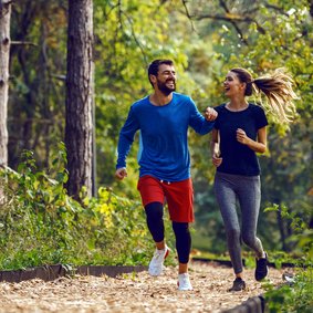 Bild: Dusan Petkovic - stock.adobe.com Zwei Personen joggen durch einen Wald und lächeln sich an.