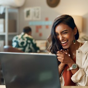 Bild: N Felix / peopleimages.com - stock.adobe.com Eine Frau sitzt vor einem Laptop und schlägt lachend die Hände zusammen.