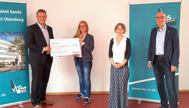 Herrmann Kruse überreicht Anita Groth einen großen weißen Spendenscheck. Daneben stehen Nadine Engel und Andreas Gögel. Bild: VHS Oldenburg.