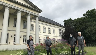 Zwei Anleiterinnen und zwei Teilnehmerinnen der Oldenburger Jugendwerkstatt stehen auf einer Rasenfläche vor einem großen Beet mit Stauden und Blühpflazen. Die Pflanzen werden zur Mitte des Beetes immer höher. Im Hintergrund steht das Gebäude des PFL. Bild: VHS Oldenburg.
