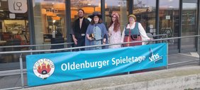 Mitglieder des Spieltraum E V in Kostümen vor dem Eingang der VHS mit einem Transparent das auf die Spieletage aufmerksam macht.