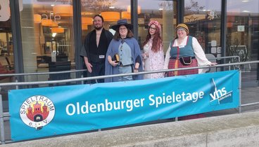 Mitglieder des Spieltraum E V in Kostümen vor dem Eingang der VHS mit einem Transparent das auf die Spieletage aufmerksam macht.