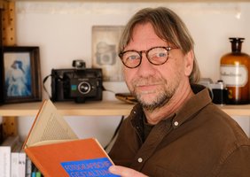 Thomas W Salzmann vor einem Regal mit Kamera und Fotos lesend in einem Buch.