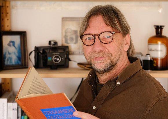 Bild: Privat Thomas W Salzmann vor einem Regal mit Kamera und Fotos lesend in einem Buch.