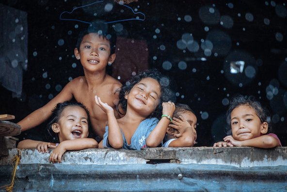 5 Kinder lehnen an einer Fensterbank und schauen zum Fotografen. Auf der Linse sind Regentropfen. Bild: Regenzeit von Hartmut Schwarzbach. 