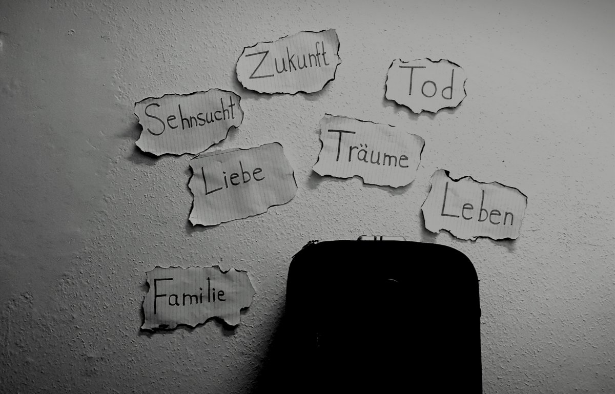 „Erinnerungen“: Die Schwarz-weiß-Fotografie zeigt einen Koffer hinter dem an der Wand verschiedene Schlagworte zu auf Papier zu lesen sind: Sehnsucht. Zukunft. Tod. Leben. Träume. Liebe. Familie. Bild: Dorgham Obaid.