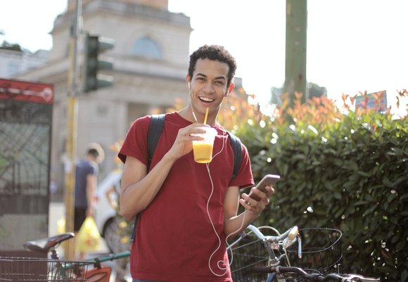 Ein Mann läuft mit Rucksack durch die Stadt und lächelt in die Kamera. In den Händen hält er ein Getränk und sein Smartphone.