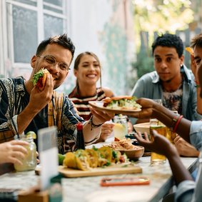 Bild: Drazen - stock.adobe.com Eine Gruppe sitzt draußen um einen Esstisch mit Tacos. Ein Mann beißt in einen Taco und reicht einen Teller weiter.