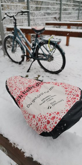 Bild: VHS Oldenburg Zwei Fahrräder an den Fahradständern der VHS im Schnee. Auf den Sätteln sind Fahrradschonern mit der Hilfetelefonnummer für Frauen.