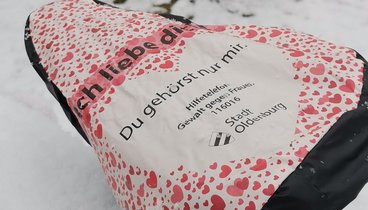 Zwei Fahrräder an den Fahradständern der VHS im Schnee. Auf den Sätteln sind Fahrradschonern mit der Hilfetelefonnummer für Frauen.