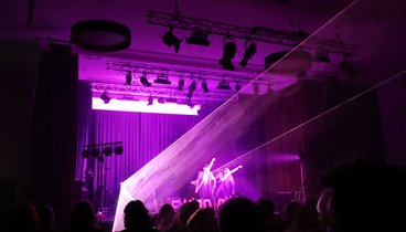 Die Protagonisten auf der Bühne in lilanem Laserlicht. Im Vordergrund die schwarzen Umrisse des Publikums.