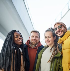 Vier Menschen unterscheidlicher Hautfarbe halten sich in den Armen und grinsen in die Kamera.