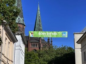 Ein Banner mit der Aufschrift "Gönn dir was" hängt zwischen zwei Gebäuden in der Oldenburg City.