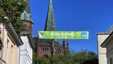 Ein Banner mit der Aufschrift "Gönn dir was" hängt zwischen zwei Gebäuden in der Oldenburg City.