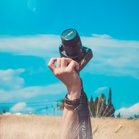 Ein Arm ragt aus einem Kornfeld und hält eine Kamera in den strahlend blauen Himmel.