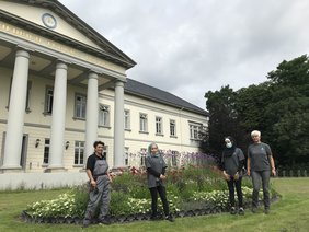 Zwei Anleiterinnen und zwei Teilnehmerinnen der Oldenburger Jugendwerkstatt stehen auf einer Rasenfläche vor einem großen Beet mit Stauden und Blühpflazen. Die Pflanzen werden zur Mitte des Beetes immer höher. Im Hintergrund steht das Gebäude des PFL. Bild: VHS Oldenburg.