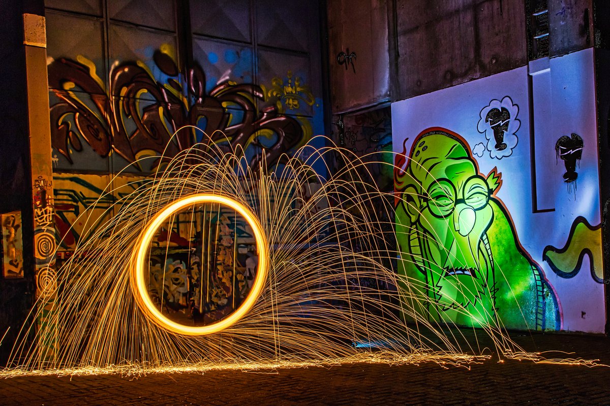 Ein Ring aus Licht (Lightpainting) sprüht im Vordergrund Funken und beleuchtet die im Dunkeln verborgenen Graffitis im Hintergrund. Die Urheber*innen der Graffiti am Utkiek, Eidechsenstraße, sind unbekannt. Bild: Klaus-Peter Kostian.  