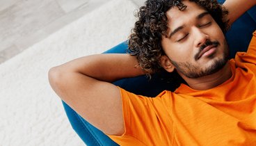Ein junger Mann im liegt mit geschlossenen Augen und den Armen hinter seinem Kopf verschränkt auf einem blauen Sofa. Er trägt ein orangenes T-Shirt. Er hat dunkle lockige Haare, einen leichten Bart und braune Haut.