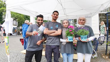 Zwei Teilnehmer und zwei Teilnehmerinnen der Oldenburger Jugendwerkstatt vor dem Stand der Jugendwerkstatt halten Blumen in die Kamera und lächeln. Bild: VHS Oldenburg.