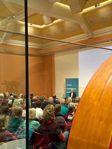 Bild: VHS Oldenburg Blick durch den Saal entlang einer alten Fleischverarbeitungsmaschine über das Publikum. Im Hintergrund sitzt Michel Abdollahi auf der Bühne.