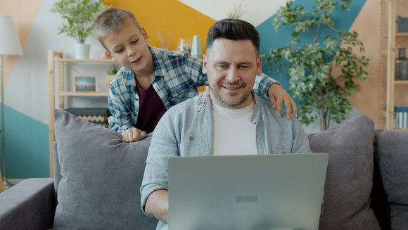 Ein Mann sitzt auf einem Sofa und shcaut in einen Laptop. Ein junge schaut ihm über die Schulter.