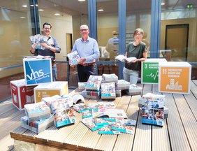 Im Lichthof der VHS liegen stapelweise Programmhefte. Veit Eveslage, Andreas Gögel und Frauke Sterwerf stehen dahinter und halten Programmhefte in den Händen. Bild: VHS Oldenburg.
