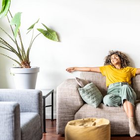 Bild: Davide Angelini - stock.adobe.com Eine Frau sitzt zurückgelehnt auf dem Sofa un breitet lachend die Arme auf der Rückenlehne aus.