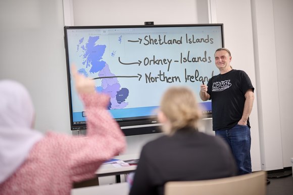 Eine Lehrkraft im Gespräch mit einer Schülerin im Unterricht. Das Smartboard zeigt Inseln vor England.