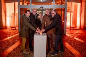 Bild: GSG Oldenburg VHS-Geschäftsführer Andreas Gögel und Vertreterinnen und Vertreter der beteiligten Insitutionen stehen im Halbkreis und drücken gemeinsam einen symbolischen orangenen Knopf, mit dem sie die Beleuchtung auslösen. Im Hintergrund der orangen beleuchtete Eingangsbereich der GSG.