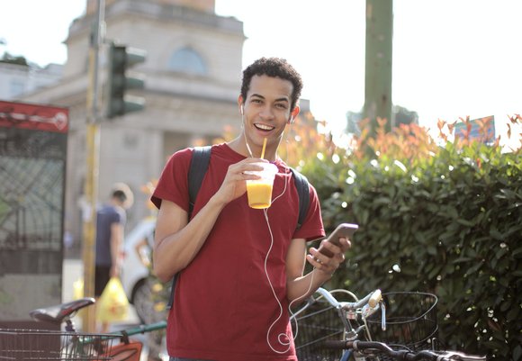 Ein junger Mann läuft mit Rucksack durch die Stadt und lächelt in die Kamera. In den Hängen hält er ein Getränk und sein Smartphone. 